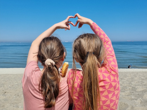 Zdjęcie pokazujące jak dwie dziewczynki siedzą obok siebie na plaży i składają swoje dłonie w kształt serca.